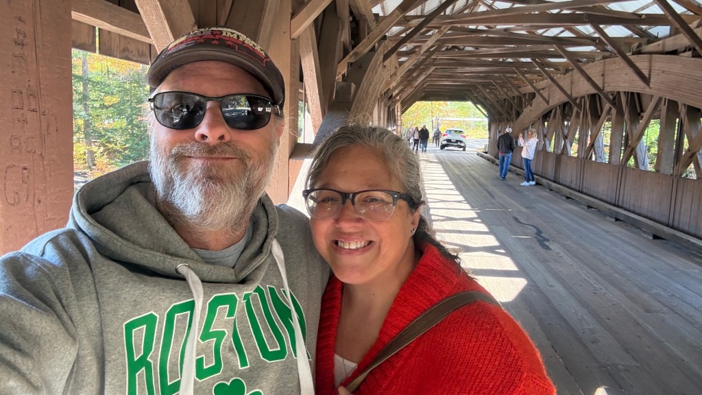 A couple smiling for a selfie inside a wooden covered bridge, with other people visible walking in the background and colorful autumn foliage outside.