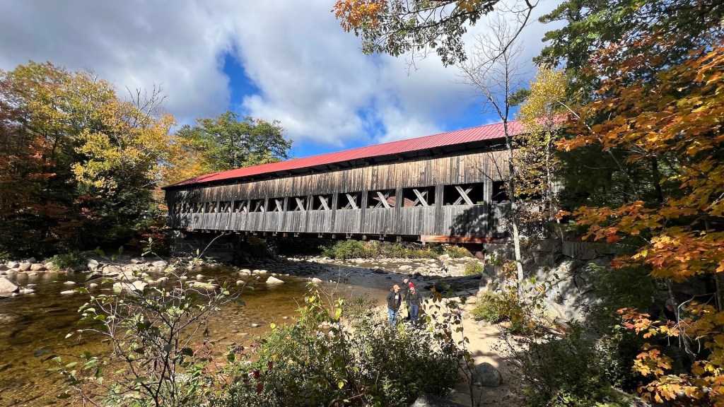 A scenic view of a covered bridge surrounded by autumn foliage, with a clear blue sky in the background and people walking near the water.