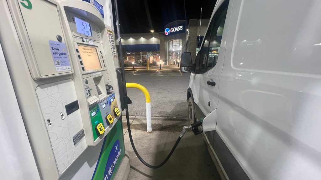 A gas pump at night with a fuel nozzle inserted into a white delivery van, and a convenience store in the background.