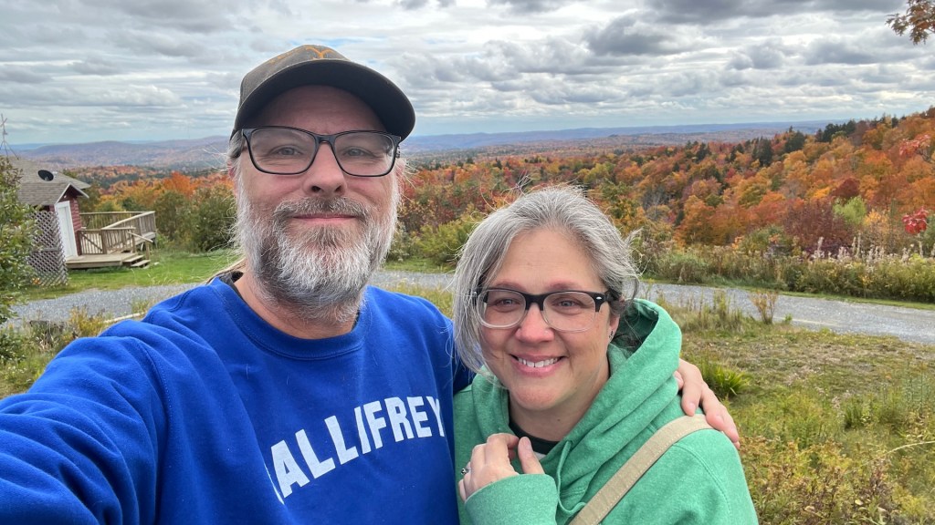 A couple smiling for a selfie against a backdrop of vibrant autumn foliage and a cloudy sky.