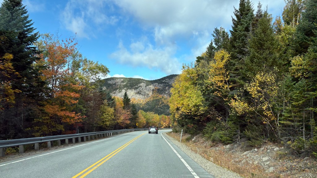 A winding road surrounded by colorful autumn foliage, with a car driving towards a mountainous background under a partly cloudy sky.