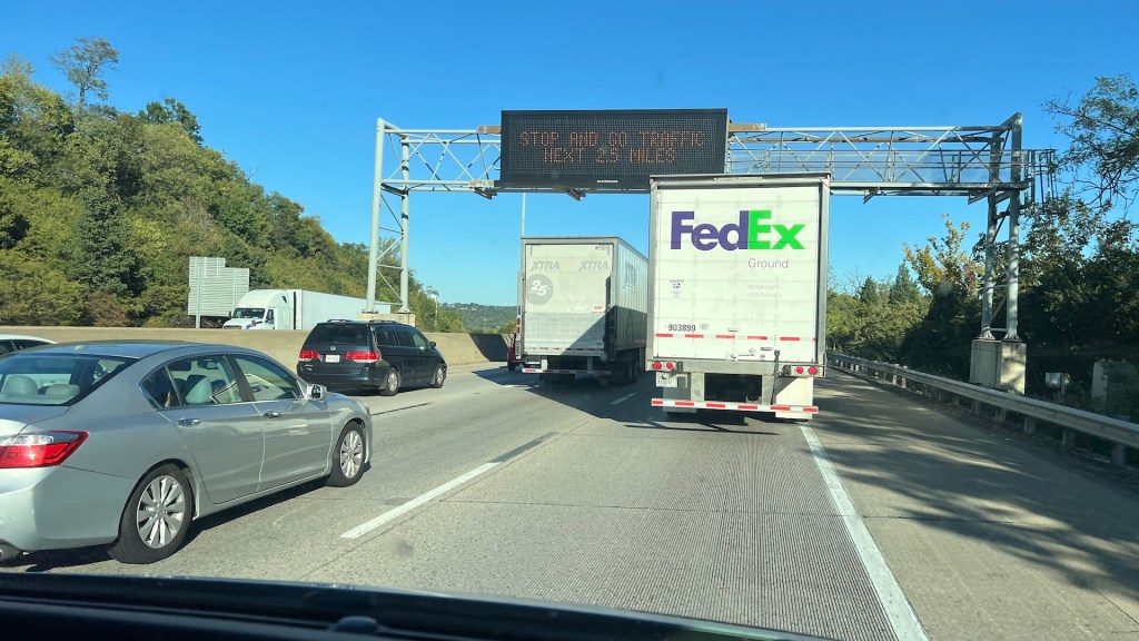Traffic on a highway with a digital sign displaying 'STOP AHEAD 1 MILE'. Several cars, including a FedEx truck, are visible.