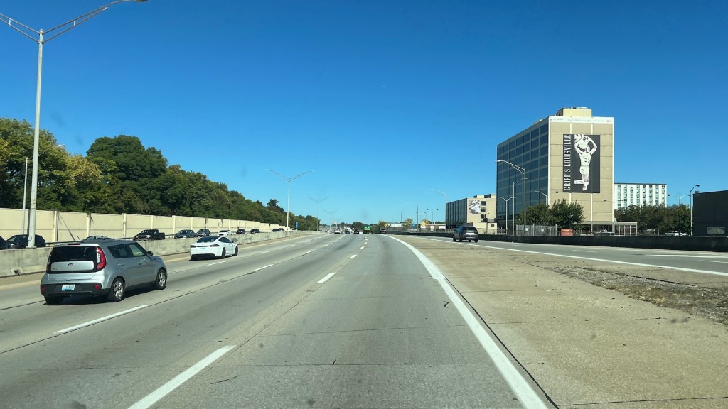 View of a highway with multiple vehicles and trees along the side, featuring a large building with a mural on its facade under a clear blue sky.