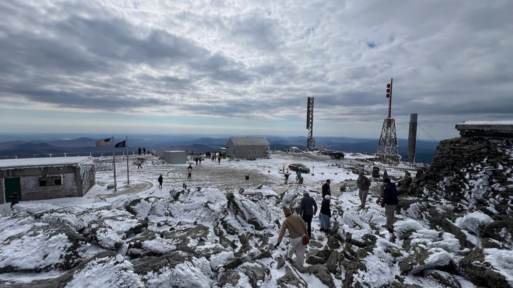 People walking on a snowy mountain summit with structures and communication towers in the background under a cloudy sky.