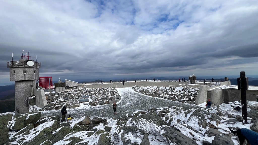 A panoramic view of a rocky mountain summit with snow cover, featuring several visitors exploring the area, a lookout tower, and cloudy skies.