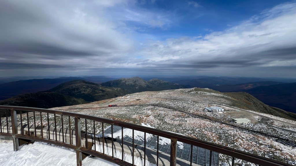 Panoramic view from a mountain peak showing a snow-covered landscape, rolling hills, and dramatic cloud formations in the sky.