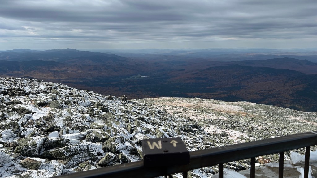 A panoramic view from a rocky mountain summit overlooking a vast landscape of rolling hills and valleys, with patches of autumn foliage visible. In the foreground, a sign indicates the direction west. The sky is cloudy, and some areas are dusted with snow.