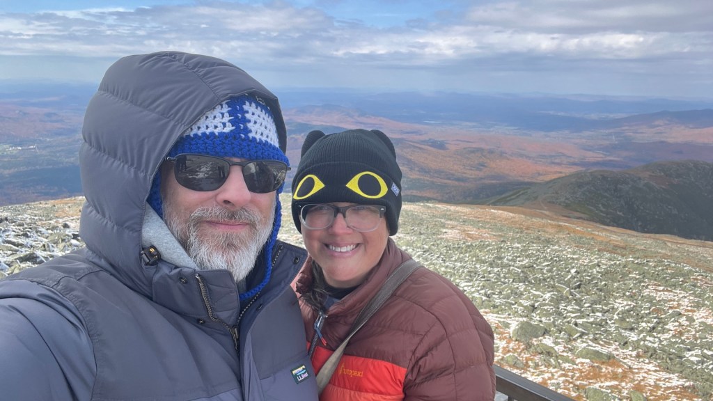 A couple posing for a selfie at a mountain summit, wearing winter clothing. The landscape features rolling hills with autumn colors and a cloudy sky in the background.