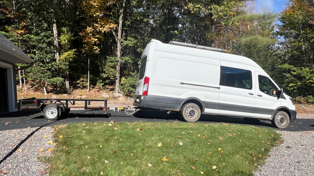A white van towing a small black trailer, parked on a driveway surrounded by green grass and trees in autumn foliage.